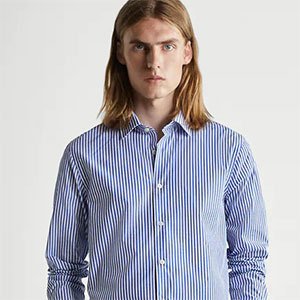 Pessoa com cabelos longos usando camisa social listrada em azul e branco, posando em frente a um fundo neutro em estilo editorial de moda.