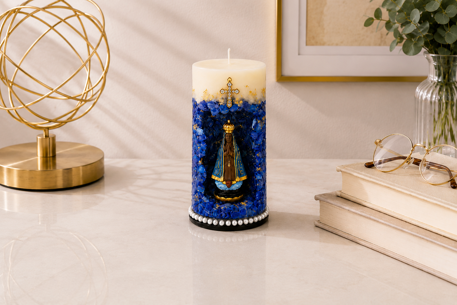 Vela Gruta de Nossa Senhora Aparecida artesanal em tons de azul profundo, branco e dourado, com imagem de Nossa Senhora Aparecida posicionada em um nicho central decorativo. A peça possui acabamento com cristais azuis, detalhes dourados, cruz ornamentada aplicada na parte superior e base adornada com pérolas. Está apresentada em ambiente sofisticado sobre mesa clara, acompanhada por livros, objeto decorativo dourado e vaso com folhagens.